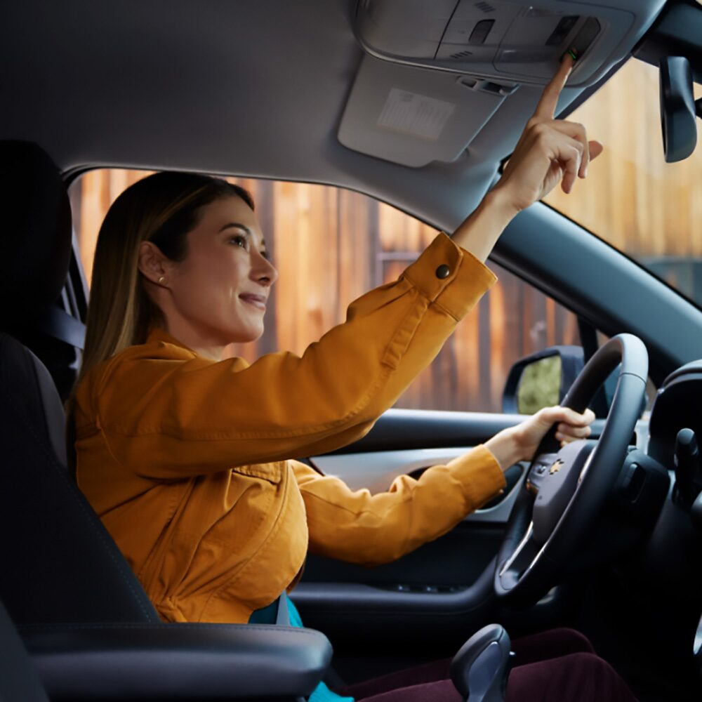 Imagen de una mujer manejando un coche Chevroley mientras utiliza la tecnología OnStar, presionando el botón ubicado frente al espejo retrovisor central. 